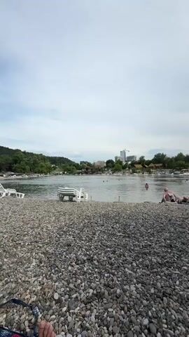 This video shows a pebble beach with several lounge chairs and thatched umbrellas. In the background, there is a body of water with some people swimming and a distant shoreline with buildings and trees. The sky is overcast.