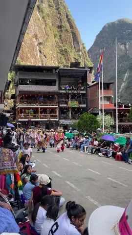 A street festival is underway in a mountainous, possibly South American, town. The street is lined with multi-story buildings, some of which appear to be restaurants or shops with balconies. On the right side of the street, people are seated on benches, watching a procession. The procession features individuals in colorful, traditional costumes, some wearing elaborate headdresses. A few people are walking in the procession, while others appear to be dancers. The street is paved, and there are flags and umbrellas visible. The background shows steep, green mountains under a bright sky. The foreground shows a group of spectators, including women and children, sitting on the ground on the left side of the street. One person in the procession is wearing a red outfit and a tall, feathered headdress. Another person in the procession is wearing a patterned headdress. The spectators in the foreground are dressed casually.
