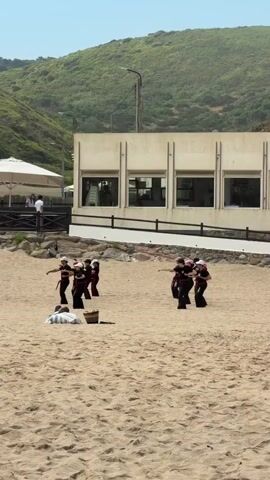 A group of individuals are practicing a synchronized movement on a sandy beach. Two groups of people are visible. One group of four individuals in matching dark outfits with white hats are arranged in a semi-circle on the sand. They appear to be performing a choreographed dance or exercise, with their arms extended in unison. In the foreground, a single individual lies on their back on the sand, appearing to be resting or observing. A brown basket sits near the person on the ground. Behind the dancing group, a modern building with large windows and a white facade stands against a backdrop of a lush green hillside. A few people are visible near an umbrella on a raised platform to the left of the building. The sky is clear and blue, suggesting a sunny day.