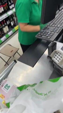 A person wearing a green polo shirt and green gloves stands behind a checkout counter in a grocery store. They are using a keyboard connected to a computer monitor. In front of them on the counter are two plastic containers of mushrooms, a wedge of cheese in plastic packaging, and a white plastic shopping bag. A payment terminal is also visible on the counter. The person's hands are moving as they operate the keyboard and handle the items.