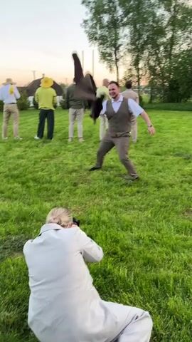 The video shows a group of men outside with a photographer, in what appears to be a rural setting. A person wearing light gray trousers and a jacket is kneeling on the grass, facing a man in a vest and trousers, and taking pictures. The man in the vest is posing, flailing his arms and jacket. There are four other men standing in the background, wearing casual attire. They appear to be observing the photo shoot. A house is seen on the left side of the video. There are some birch trees in the background. The sky is light with an orange glow at the horizon. The setting looks like an open grassy field.