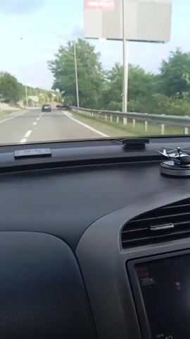 The video shows a first-person perspective from inside a car, looking out the windshield and dashboard. The car is moving on a paved road, indicated by white lines and a guardrail on the right. Trees and vegetation line the side of the road. In the distance, there are power lines and poles. A small helicopter-shaped air freshener is visible on the dashboard. At one point, a signpost and a small bus stop structure are visible by the roadside. The sky is clear and bright.