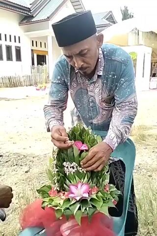 Here is a description of the video:

The video shows a man decorating a conical floral arrangement with pink flowers. The arrangement is made of green leaves and other small white flowers and sits on a blue chair wrapped in red tulle. The man wears a black taqiyah (a type of skullcap), a light blue patterned long-sleeved shirt, and a dark sarong. He carefully places the flowers into the arrangement with his hands. The setting appears to be outdoors, in front of a house, with another person walking by in the background. A white tent-like structure is also visible in the background.