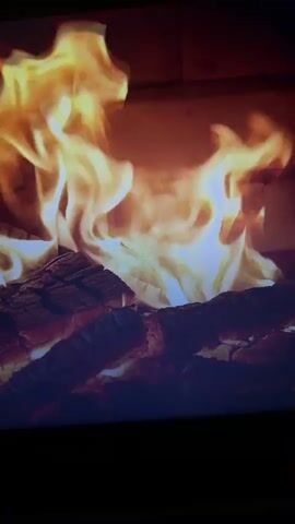 The video depicts a close-up view of a fireplace with burning logs. Flames are visible above the logs, exhibiting bright yellow and orange hues. The logs themselves appear dark and charred. The background suggests the interior of a brick fireplace. The video is relatively static, focusing primarily on the burning fire.