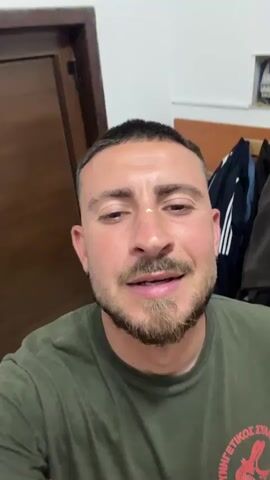 The video features a close-up, selfie-style shot of a smiling man indoors. He appears to be in his 20s or 30s, with short dark hair, a neatly trimmed beard, and brown eyes. He wears an olive green t-shirt with a partially visible red and white graphic. A small, distinct white mark is present on the bridge of his nose. He is speaking directly into the camera, with a wide, cheerful smile, indicating a positive and engaging mood.

The background features a dark wooden door on the left and a plain white wall dominating the center and right. On the right side, an open electrical fuse box with visible wires and breakers is mounted on the wall. Below the fuse box, two dark-colored jackets or hoodies, one with white stripes, hang on hooks.

The graphic on the man's t-shirt clearly displays Greek text: "ΚΥΝΗΓΕΤΙΚΟΣ ΣΥΛΛΟΓΟΣ" (Kynigetikos Syllogos, meaning "Hunting Club/Association") and "ΛΑΓΟΣ" (Lagos, meaning "Hare/Rabbit"), accompanied by a red, lobster-like emblem. The overall setting suggests a casual, possibly functional or backroom environment. The video's location context is Lympia, Cyprus, which is consistent with the Greek language on the shirt.