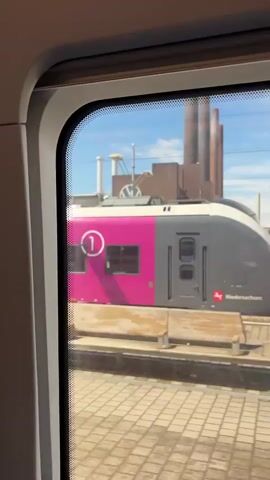 A train moves past a large industrial complex with multiple smokestacks and a prominent Volkswagen logo on the side of one of the buildings. The view is from inside the train, looking out a window. A train platform with tracks and paving stones is visible in the foreground. The sky is clear and blue.