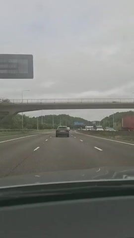 The video shows a view from inside a vehicle driving on a highway. A pedestrian bridge spans the highway in the foreground. Several cars and trucks are visible on the highway, moving in the same direction as the camera vehicle. The sky is overcast, and there are trees and hills on either side of the highway. A traffic sign is visible in the distance.