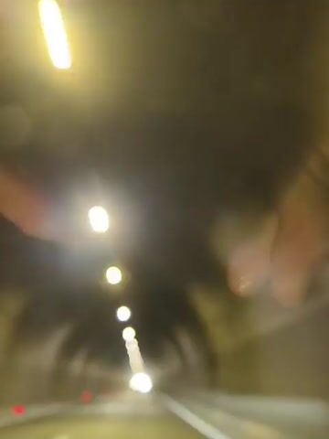 The video shows a view from inside a vehicle driving through a tunnel. The tunnel is illuminated by rows of lights on the ceiling. The walls of the tunnel appear to be made of concrete. The road ahead is visible, with lane markings. There are no people visible in the video. The perspective suggests the camera is mounted on the dashboard or windshield. There are some light flares and reflections present in the video.
