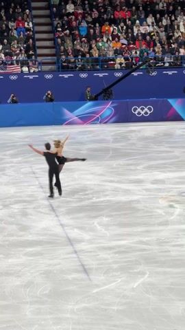 The video shows a male and female ice skating pair performing a lift on an ice rink. Both are wearing black outfits. The female has blonde hair. The rink has a bright surface with skate marks. A row of Olympic Rings decorates the rink’s wall. Spectators fill the stands in the background. A camera with a boom pole is visible near the wall. The location is branded for "MILANO CORTINA 2026." After the lift, the pair continues skating, maintaining a synchronized dance.
