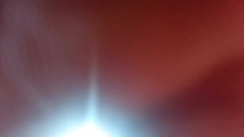 The video shows a very blurry, reddish-brown abstract image. There is a bright light source on the right side of the frame, casting a white glow that is partially obscured by a pinkish hue. The overall impression is of a close-up, out-of-focus shot, possibly of a hand or fabric, with strong backlighting. There is no discernible action or interaction, and the environment is entirely undefined.