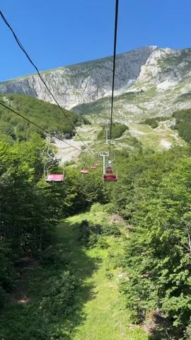 A chairlift ascends a mountainside on a clear, sunny day. Lush green trees line the lower slopes, giving way to a more sparse, rocky terrain higher up. The chairlift, with its red seats, moves up the incline, disappearing into the distance towards the mountain peak. The sky is a vibrant blue.