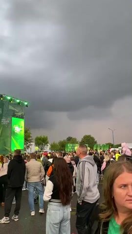 A large crowd of people are gathered outdoors, facing a stage. The stage is decorated in green and has lights. A banner on the stage reads "BELARUSBANK". The sky above is dark and cloudy, suggesting impending rain or a storm. The people are standing, some looking towards the stage, others talking amongst themselves. The overall atmosphere appears to be that of an outdoor event or concert.