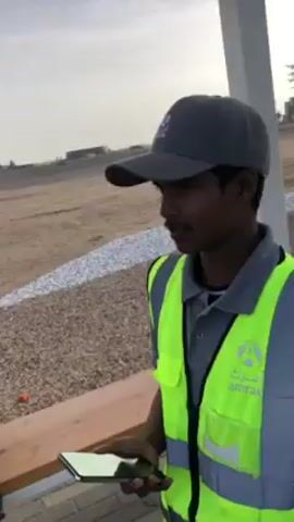 A young man stands outside, smiling, and briefly speaks to the camera. He is wearing a gray collared shirt, a bright green safety vest, and a gray baseball cap. The vest has the logo of "amrak" on the right side. He is holding a smartphone in his left hand. The background includes a light-colored brick sidewalk, a wooden bench, a beige gravel area, a light-colored building in the distance, and a tall, slender white structure.