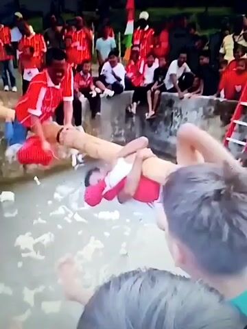 A group of people in red shirts watch as two men attempt to balance on a wooden log suspended over water. One man is partially submerged in the water, his face visible, and appears to be struggling to stay on the log. The other man is on top of the log, his hands on it, and seems to be trying to help the submerged man. The scene appears to be some kind of competition or game.