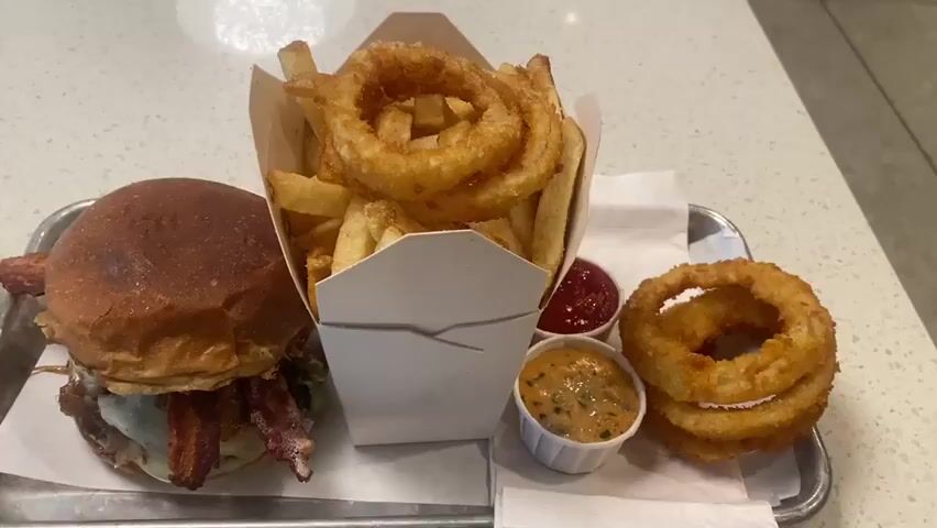 The video shows a close-up, overhead shot of a meal served on a metallic tray. The meal consists of a large hamburger, a container of french fries and onion rings, two dipping sauces, and a small stack of onion rings. The hamburger is on the left, with a toasted bun, lettuce, cheese, and bacon visible. The container of fries and onion rings is in the center, with fries forming the base and several golden-brown onion rings piled on top. To the right of the container are two small white ramekins, one filled with a red sauce (likely ketchup) and the other with a creamy, orange sauce with visible herbs. A small stack of three onion rings sits to the far right of the tray. The background is a speckled white countertop. The lighting is bright, creating some glare on the tray and food. The camera angle remains stationary throughout the short clip.