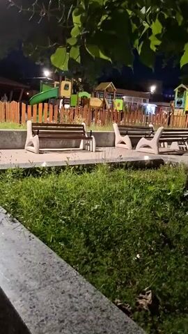 The video shows a nighttime view of a playground and park area. In the foreground, there is lush green grass and a portion of a stone ledge. Behind the grass, there are several modern, curved park benches. Further back, a playground with colorful slides and structures is visible, enclosed by a wooden fence. The area is illuminated by streetlights and lights from buildings in the background. Several people can be seen walking and standing around the park area, including a group of four individuals near one of the benches and two people on the playground. A tree with green leaves is prominent in the middle ground. The overall atmosphere appears to be a quiet, public space at night.