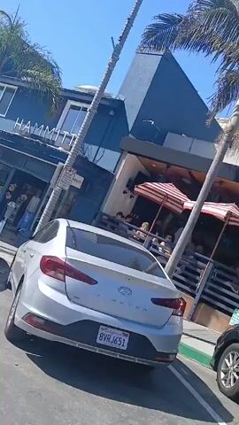 The video shows a street scene with parked cars and a building with the name "HAMMITT" displayed on its facade. Palm trees are visible in the background, and the sky is clear and blue. A silver Hyundai Elantra is parked closest to the camera, followed by a grey Volkswagen Jetta and a black Mercedes-Benz C-Class. People can be seen walking near the building, and some are seated at an outdoor cafe with red and white striped umbrellas. The camera appears to be moving slowly, possibly from inside a car. The overall environment suggests a sunny, possibly coastal, urban area.