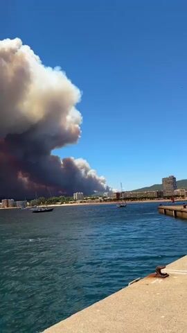 A panoramic video displays a coastal town under a sky filled with a massive, ominous plume of smoke from a wildfire. The smoke, colored in shades of white, gray, and dark red, billows dramatically from the left side of the frame, stretching across the horizon. In the foreground, the clear blue ocean water is visible, with gentle waves. A stone embankment or pier occupies the bottom right corner of the frame. Several sailboats are anchored in the bay, and a large cruise ship is docked at a pier on the right side of the screen, dwarfed by the immense smoke cloud. The town, with its buildings, is situated along the coastline, partially obscured by the smoke. The sky is a clear, bright blue, contrasting sharply with the dark smoke. The scene suggests a significant and ongoing wildfire event impacting a populated coastal area.