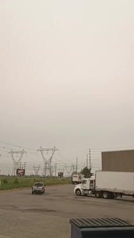 A white semi-truck with a white trailer is parked to the right of the frame. A silver pickup truck is driving away from the camera to the left of the frame. In the background, there are power lines and power towers. The sky is overcast and appears hazy.