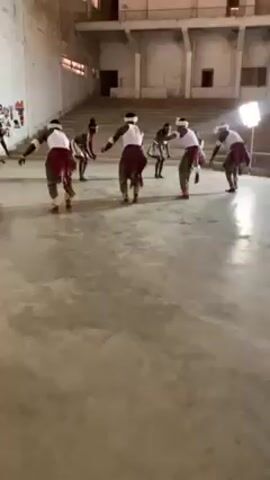 Several people are performing a dance in a large, empty auditorium. The dancers are wearing white tops, dark bottoms, and light-colored leg coverings. They also have headbands and adornments around their waists. The dance involves synchronized movements, including bending over and stepping. The setting appears to be an old, somewhat run-down hall with concrete floors and tiered seating in the background. There is a lighting stand on the right and what appear to be decorations on the left.