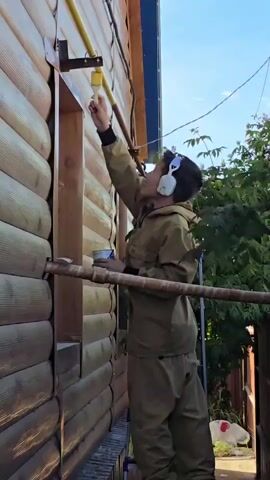 A person in a tan jumpsuit and headphones is standing on a ladder and painting a yellow pipe attached to the side of a wooden house. The person is holding a paintbrush and a can of paint. The house is made of logs and has a window. There are trees and bushes in the background. The sky is blue with some clouds.