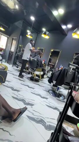 The video shows the interior of a barbershop.  The floor is tiled with a swirly marble pattern in white and grey. There are three barber chairs, each with gold and black details. Two are occupied by customers getting haircuts. One man is getting a haircut from a barber dressed in a white shirt and black pants. Another barber with blonde hair is standing behind the customer and the barber. The barbershop has mirrors along one wall and stylish light fixtures hanging from the ceiling. A trolley with barber supplies is near the barber chair in the foreground. In the bottom left corner, a person’s crossed legs are visible, showing a foot wearing a black sandal. Music is playing in the background.