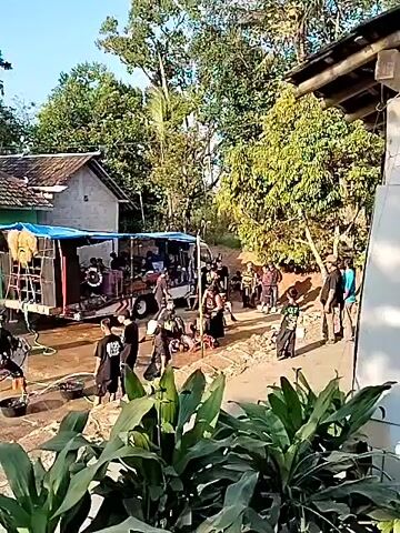 The video shows an outdoor gathering in a rural setting, likely a performance or cultural event. In the foreground, lush green plants partially obscure the view. The scene unfolds in a courtyard area in front of traditional houses. Several people are gathered, some seated on the ground and others standing. In the center of the courtyard, individuals in traditional attire appear to be performing, with at least two individuals in what looks like animalistic costumes, possibly a horse or lion dance. A trailer with speakers and equipment is set up to the left, suggesting amplified music or sound for the performance. Other individuals, possibly performers or organizers, are seen around the trailer and in the background. Further back, more people are watching the performance, creating a sense of community engagement. The environment is characterized by simple housing, trees, and a dirt or gravel ground. The lighting suggests it is daytime, likely morning or afternoon, with sunlight casting shadows. The overall atmosphere is one of a lively, local event.