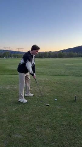 The video shows a young man playing golf on a green field. He is wearing a dark sweater with a light-colored X design, beige pants, and light-colored shoes. He swings a golf club and hits a golf ball, then watches the ball as it flies into the distance. Behind him, there is a wide open green field with some trees in the background, as well as mountains and a clear sky. The sun is setting, casting a golden glow on the horizon. There are power lines visible in the distance. A black golf tee sits in the grass next to the golf ball.