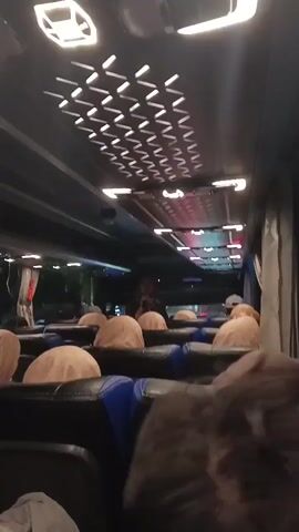 The video shows the interior of a bus at night. The camera is positioned facing forward, looking down the aisle. Rows of empty, beige-covered seats are visible on either side. The ceiling of the bus features modern, angular lighting fixtures, some of which are illuminated. In the distance, a person can be seen standing in the aisle. The overall lighting is dim, with some red and blue lights visible further down the bus. The view suggests that the bus is either stationary or moving slowly.