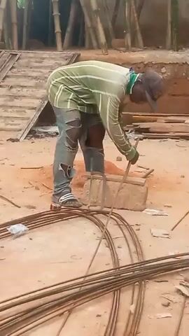 A construction worker is depicted actively bending rebar at a construction site in Ihiala, Nigeria. The man, wearing a light green and white striped long-sleeved shirt, dark blue ripped jeans with visible tears, black and white striped slide sandals, a dark baseball cap, and work gloves, is deeply focused on his task. He is bent over a metal bending jig, which is stably placed on a concrete block, using it to shape a piece of steel reinforcing bar.

Surrounding him on the reddish-brown dirt ground are numerous lengths of rebar, some coiled and some straight, indicating ongoing construction work. In the background, elements of the construction site are visible, including bamboo scaffolding and scattered wooden planks. A small mound of excavated earth is also present behind the worker. The scene is brightly lit by natural daylight, suggesting a clear day. No visible text is present.