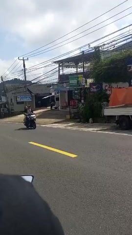 The video shows a view from a moving motorcycle on a road. The road is paved and has yellow lane markings. The video passes by a series of buildings including a shop that advertises "JUAL ES BATU KRISTAL". There are many power lines overhead. There are several vehicles including cars and motorcycles. One motorcycle has two people riding on it. There is a person standing in front of a white van near a shop. The sky is partly cloudy.