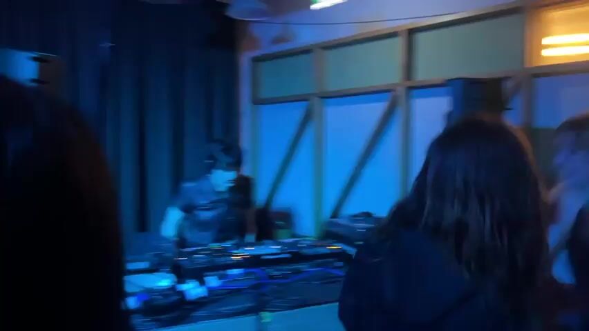 The video captures a DJ performing a set in an indoor club or event setting in Dundee, United Kingdom. A male DJ with dark hair, wearing headphones and a black leather-like short-sleeved top, is seen actively mixing music at a DJ booth. His setup prominently features a "Pioneer DJ" mixer, flanked by two turntables, and an array of cables. Drinks in glasses are also visible on the DJ table. A large black speaker stands to the left of the booth. The room is bathed in a distinct blue light, illuminating blue curtains in the background and wooden structural elements. Several individuals, mostly seen from behind with long dark hair, are in the foreground, observing the DJ and enjoying the music. The atmosphere suggests an evening or nighttime event, with the DJ focused on his equipment and the audience engaged in the performance.