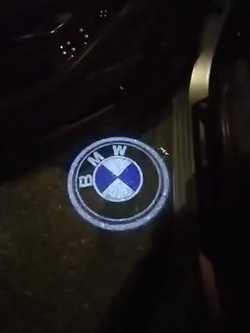 The video shows a close-up view of a BMW logo projected onto the ground from a car door. The logo is circular with the letters BMW in white around a blue and white cross shape in the center. The projection is illuminated and appears to be a door sill light. The background is dark, suggesting it is nighttime or the inside of a dimly lit car.