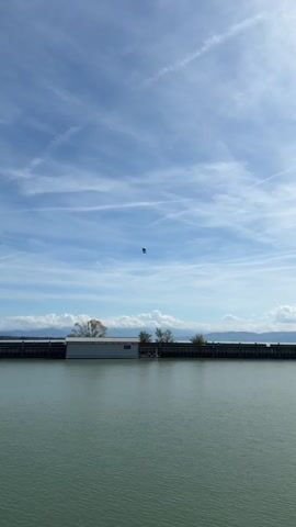 The video shows a zeppelin flying in the sky over a body of water. In the foreground, the water is calm and a greenish-blue color. In the background, there is a long, low building with a white roof and gray walls, which appears to be a boathouse or dock. Trees are visible on either side of the building. Beyond the building, there is a railing or pier that extends along the water's edge. In the distance, there are clouds and mountains. The zeppelin is a silver and blue airship with a dark gondola underneath. It is the sole focus of the camera's attention as the video progresses, with the view zooming in on the zeppelin as it flies higher into the sky. The sky is blue with scattered white clouds.
