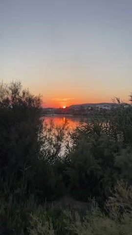 The video shows a sunset over a body of water. The sun is low on the horizon, casting a warm orange glow on the water and the sky. There are trees and vegetation in the foreground, framing the scene. In the distance, there appear to be some buildings or structures on a hill. The water is calm, with a reflection of the sunset visible on its surface. The overall mood of the video is peaceful and serene.