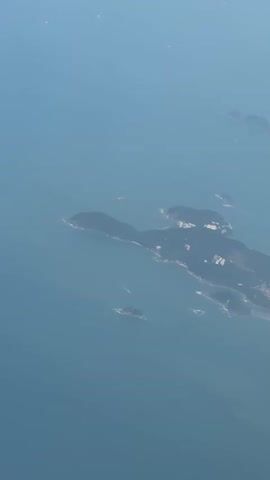 A high-angle aerial view from an airplane shows a vast expanse of blue ocean and a coastal cityscape. The city, characterized by numerous tall buildings and a long stretch of sandy beach, runs along the shoreline. Several smaller islands are scattered in the surrounding ocean. The sunlight illuminates the scene, creating shadows on the buildings and landmasses. The camera slowly pans to the right, revealing more of the coastline and the distant city. The clear blue sky is visible at the top of the frame.