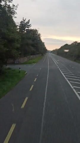 The video shows a view from a vehicle traveling on a paved road. The road has two lanes in the direction of travel, indicated by white dashed lines. The left side of the road is bordered by a stone wall and lush green grass, with a row of trees lining the wall. On the right side, there are also trees and vegetation. The sky is overcast with hints of orange and yellow light from what appears to be a sunrise or sunset. There are other vehicles visible in the distance on the road. The overall impression is a scenic drive through a rural or suburban area during a period of transitional light.