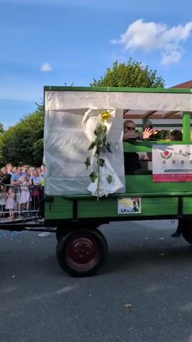 A video shows a green trailer with people inside and a man in an orange vest walking beside it. The trailer is decorated with white plastic sheeting on the sides and a large sunflower in the front. A banner on the side of the trailer reads "IlmtalCare" with a heart logo. People are sitting and standing inside the trailer, some of whom are looking towards the camera. The man in the orange vest is holding a blue object in his hand and looking towards the trailer. The video is filmed outdoors during the daytime, with a blue sky and green trees visible in the background. There are other people in the background, suggesting a public event or parade.