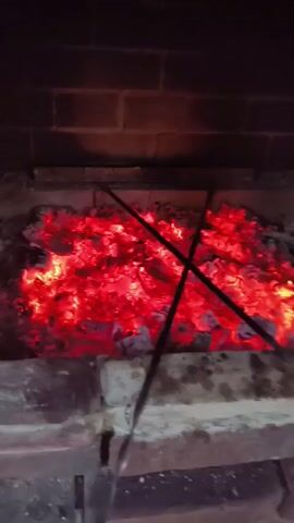The video shows an overhead view of a fireplace filled with glowing red embers. Two long metal skewers are placed across the top of the embers, angled towards each other in an X formation. The background consists of dark brickwork. The embers appear to be hot and are radiating heat. The scene is dimly lit, with the primary light source coming from the glowing embers.