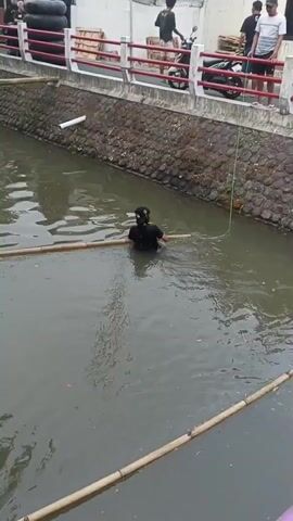 A person wearing a black t-shirt and a black baseball cap is standing in murky water, holding onto a bamboo pole that stretches across the width of the waterway. The person appears to be wading or trying to navigate the water. Another bamboo pole is visible in the foreground, also spanning the water. The waterway has stone retaining walls on one side and what appears to be a concrete walkway or bridge above. A thin green rope is also visible in the water, attached to something out of frame on the right.