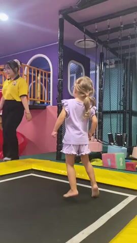 A young Caucasian girl with blonde hair in a ponytail, wearing a light purple t-shirt and shorts, is jumping on a trampoline. The trampoline has a black jumping surface and a yellow padded frame. The background shows a colorful indoor playground with purple walls, a red slide, and a ball pit filled with pink, blue, and white balls. There are also some large, colorful blocks with numbers on them visible. The girl appears to be mid-air, with her feet off the ground, and she is looking to her left. The lighting is bright and appears to be artificial.