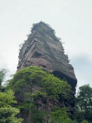 The video shows a very tall, imposing rock formation in a natural environment. The rock is primarily gray and red, with visible layers and stratification. It is partially covered with greenery, including trees and shrubs at its base and some vegetation on its upper parts. The sky is overcast, contributing to the muted, slightly hazy atmosphere. Sounds of people talking can be heard, suggesting the presence of onlookers or tourists nearby. The camera angle is low, looking upwards, which emphasizes the height and scale of the rock formation.
