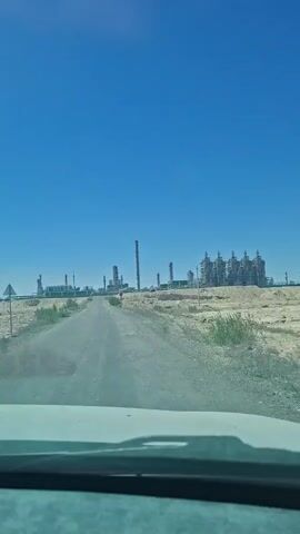 A video shows a view from inside a vehicle driving on a dirt road toward an industrial facility under a clear blue sky. The road is dusty and uneven, with sparse vegetation on either side. In the distance, a large complex of industrial structures, including towers and interconnected buildings, is visible. The facility appears to be an oil refinery or a similar industrial plant. The sun is bright, casting shadows. The video is shot from a low angle, likely from the dashboard of the vehicle. There are no people or animals visible in the frame. The overall scene suggests an industrial landscape in an arid or semi-arid environment.