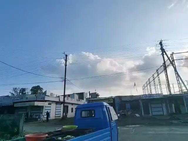 The video shows a street scene with several small buildings, some of which appear to be shops, lining the road. A blue three-wheeled vehicle is parked on the left side of the frame. In the background, there is a large fenced enclosure, possibly a sports court, with the words "THE REAL TURF" visible on a sign. There are several power lines overhead, supported by utility poles. The sky is blue with scattered white clouds, suggesting it is daytime. Some people are visible near the shops and the sports facility, and a few motorcycles and a car are parked.