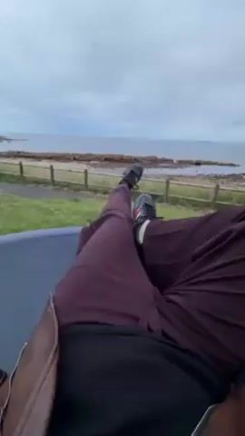 Here is a concise description of the video:

The video shows a person lying on a swing, presumably a basket swing, overlooking a sea or ocean view. The person is dressed in brown pants, black shoes, and a black top. They are wearing a brown leather jacket and briefly come into view while lying back. They make a peace sign, then close their eyes and remain still. The swing is on a grassy area near a fence, with a beach and water visible in the background under a cloudy sky.