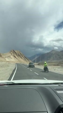 The video shows a first-person view from inside a vehicle driving on a paved road through a mountainous, arid landscape. The road is asphalt with white dashed lines marking the lanes. On the left side of the road are rugged, light brown mountains. On the right side, there is a dry riverbed or gully with rocks and sparse vegetation, and beyond that, more mountains. Utility poles with electrical wires are visible along the roadside. The sky is partly cloudy with dramatic formations of grey and white clouds, suggesting a potentially changing weather. A dark-colored SUV is visible driving ahead on the same road. The interior of the vehicle is partially visible in the foreground, including the dashboard and a portion of the windshield with some prayer flags attached. The overall environment appears to be a remote and scenic route, possibly in a high-altitude region.