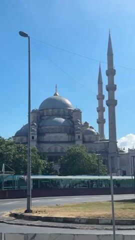 The video captures a view of a large mosque with multiple domes and minarets against a clear blue sky. In the foreground, there is a street with a light pole and some greenery. A bus is visible in the middle ground, and further in the distance, a cityscape with more buildings and trees can be seen. The video pans slightly to the right, revealing more of the mosque and its surroundings. The overall atmosphere is bright and sunny.
