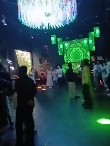 The video depicts a nightclub or event space with a diverse crowd of people. Some are standing, chatting, and milling around, while others are seated. The lighting is dim, creating a typical nightlife atmosphere, with colored lights and a large, illuminated, cylindrical chandelier hanging from the ceiling. The walls are decorated with large, screen-like panels displaying abstract green designs. The scene appears to be a social gathering or party, with people dressed in a variety of casual and semi-formal attire.