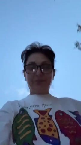 This is a first-person perspective video clip of a woman wearing glasses and a white t-shirt with a colorful fish design and the text "MUNDO MAYA, MEXICO" printed on it. She appears to be outdoors during dusk, as indicated by the blue, cloudy sky and the lighting. The background shows parts of buildings and some foliage, suggesting a suburban or rural setting. The woman is looking directly at the camera and has a neutral expression. The video is brief and appears to be a selfie taken by the subject.