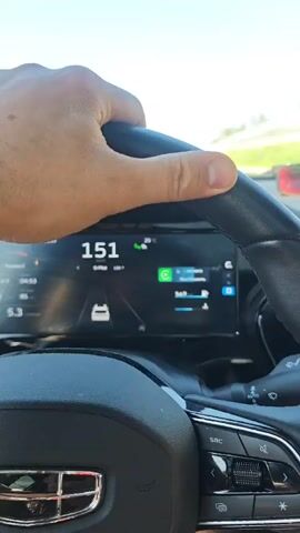 A person is driving a car on a sunny day, based on the clear sky visible through the windshield. The dashboard displays a digital speedometer, which shows the car’s speed accelerating from 152 to 164, kilometers per hour. A navigation system is visible on the digital dashboard. The driver's hand is gripping the black leather steering wheel. The car is equipped with various buttons and controls on the steering wheel. The environment appears to be a highway, with green fields and other vehicles visible in the distance. Music is playing in the background.